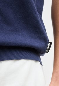 Κοντινό πλάνο ενός ατόμου που φοράει μπλε πλεκτό πουλόβερ με ετικέτα της μάρκας ECOALF και άσπρο παντελόνι, δείχνοντας την υφή του υφάσματος και τις λεπτομέρειες της ραφής.