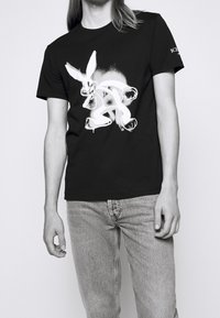 Sort bomulds T-shirt med grafisk hvid tegning af en tegneseriekanin. Rund halsdesign, korte ærmer og afslappet pasform. Lyse denim jeans synlige.