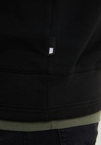 Felpa nera con texture a costine, caratterizzata da un piccolo etichetta nera con un rettangolo bianco, sovrapposta a un orlo di maglione verde.