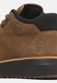 Brązowy skórzany but z czarnym materiałowym kołnierzem, metką GORE-TEX, emblematem wodoodpornym i teksturowaną czarną kauczukową podeszwą. Posiada beżowe przeszycia.