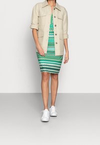 Kvinna klädd i en grönmönstrad knälång klänning, beigt knappjacka med uppvikta ärmar och vita sneakers framför en neutral bakgrund.