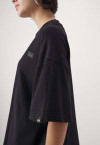 Camiseta oversized de algodón negra con mangas cortas, que presenta un parche de logo texturizado en el pecho y un pequeño detalle dorado en el dobladillo.