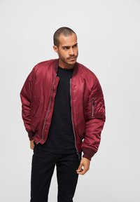 Veste bomber bordeaux avec une finition lisse et brillante, des poignets et un ourlet côtelés, une poche zippée sur la manche, portée sur un t-shirt noir.