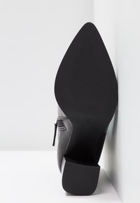Semelle de bottine noire à bout pointu avec grip texturé et talon bloc, photographiée en position verticale contre un mur blanc et un sol blanc.