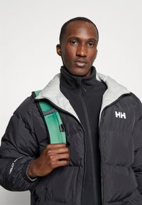 Zwarte pufferjack met grijze kraag en "HH" logo, gelaagd over een zwart zip-up top. Een groene rugzakriem is zichtbaar op de schouder.