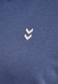 Deux chevrons blancs brodés sur un tissu bleu texturé, centrés dans le cadre.