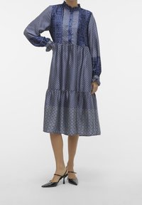 Abito blu navy in tessuto stampato, con maniche lunghe, chiusura frontale con bottoni e un orlo a strati. Tacchi neri a punta completano l'outfit.