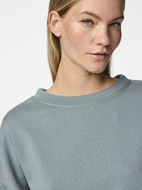Nærbilde av en kvinne med blondt hår som har på seg en lyseblå crewneck-genser mot en enkel hvit bakgrunn.