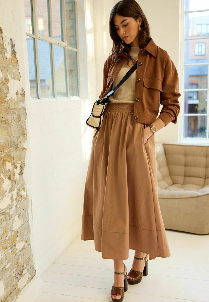 Brun jakke, beige top og lang, flydende tan nederdel. Strappy brune hæle og en lille tofarvet skuldertaske. Struktureret væg og lys indretning.