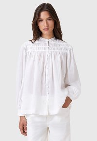 Blusa bianca leggera a maniche lunghe con colletto a volant, chiusura frontale con bottoni e dettagli arricciati alle spalle e al petto.