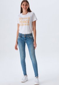 Camiseta blanca con texto amarillo, combinada con pantalones ajustados azul claro. El conjunto se complementa con zapatillas blancas.