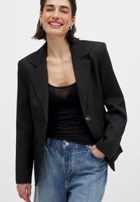 Svart skräddarsydd blazer med skarpa slag och en enda knappstängning, bärs över en svart linne, ihop med ljusblå denimjeans.