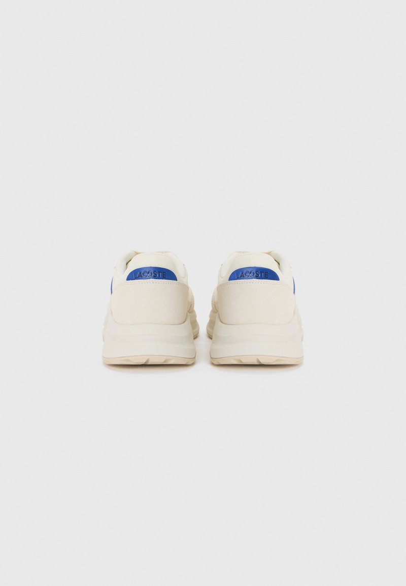 Une paire de baskets beiges Lacoste avec des patchs de logo bleus sur les talons, montrées de l'arrière sur un fond blanc.