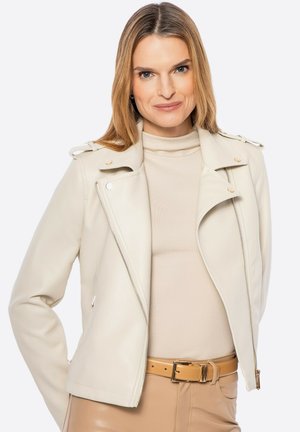 Beige neplederen motorjack met zilverkleurige hardware, asymmetrische voorknoopsluiting en schouderepauletten, gedragen over een crèmekleurige coltrui.