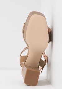 Sandale en daim beige avec talon bloc, bride à la cheville et bout carré, vue du côté semelle sur fond blanc.