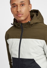 Green, white, and navy color-blocked jacket with a zip front, adjustable hood, and smooth texture, featuring a slight sheen and stitched detailing.