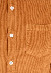 Camicia in velluto a coste marrone con una superficie testurizzata, taschino sul petto e bottoni bianchi sul davanti. Presenta un motivo a costine verticali.