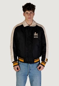 Giacca bomber nera in satin con maniche crema, colletto e orlo a coste con strisce gialle. Include il logo "BOSS" sul petto.
