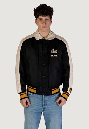 Chaqueta bomber de satén negra con mangas crema, cuello y dobladillo de canalé con rayas amarillas. Incluye el logo "BOSS" en el pecho.