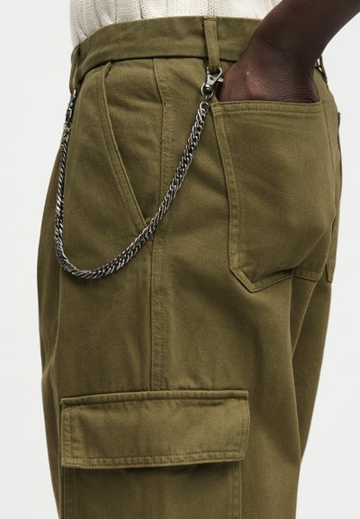 Σκούροχρωμο χέρι στην τσέπη ελιάς παντελονιού cargo με ενσωματωμένη ασημένια αλυσίδα που κλιπάρει στον θηλιά ζώνης και στην άκρη της εμπρόσθιας τσέπης.