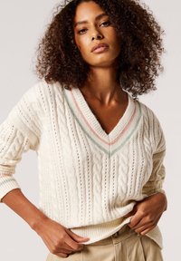 Cremfarbener Strickpullover mit V-Ausschnitt. Besitzt pastellfarbene Streifen in Rosa und Grün entlang des Ausschnitts. Strukturiert, mit langen Ärmeln.