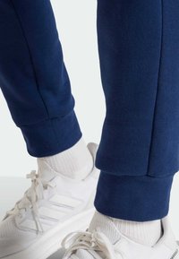 Ξεθωριασμένο μπλε φόρμα παντελόνι με σφηνοειδές σχέδιο, με ριμπ μανσέτες. Συνδυασμένο με λευκά αθλητικά παπούτσια με τρεις ρίγες και λευκές κάλτσες.