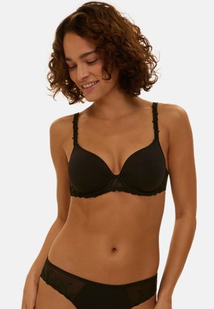 Vrouw met krullend haar, gekleed in een zwarte beha met kanten afwerking en bijpassend ondergoed, glimlachend en naar beneden kijkend met een neutrale achtergrond.