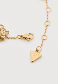 Χρυσό αλυσίδα βραχιόλι με λουλουδάτο γούστο που διαθέτει διαυγείς πέτρες, κλείσιμο τύπου αστακού και τριγωνική ετικέτα χαραγμένη με το "GUESS."