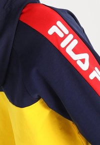 Giacca blu navy con metà inferiore gialla, con una fascia rossa sulla spalla e logo "FILA" bianco. Tessuto morbido con design sportivo.