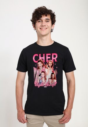 Smilende ung mand iført sort T-shirt med pink "CHER" tekst og fire kvindelige figurer med sloganet "Ugh! As if!" på fronten.
