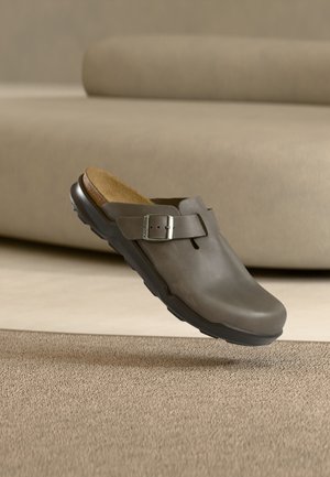 Chaussure en cuir marron à enfiler avec boucle réglable et semelle intérieure en liège rembourrée sur tapis beige et fond de canapé.