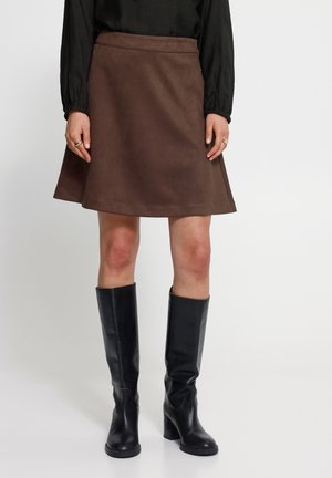 Femme portant une jupe marron mi-genou, un haut noir à manches longues et des bottes noires montantes à talons, debout sur un fond blanc.