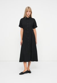 Donna in piedi con indosso un vestito midi nero a maniche corte con bottoni e colletto, abbinato a ballerine nere, contro uno sfondo bianco uniforme.