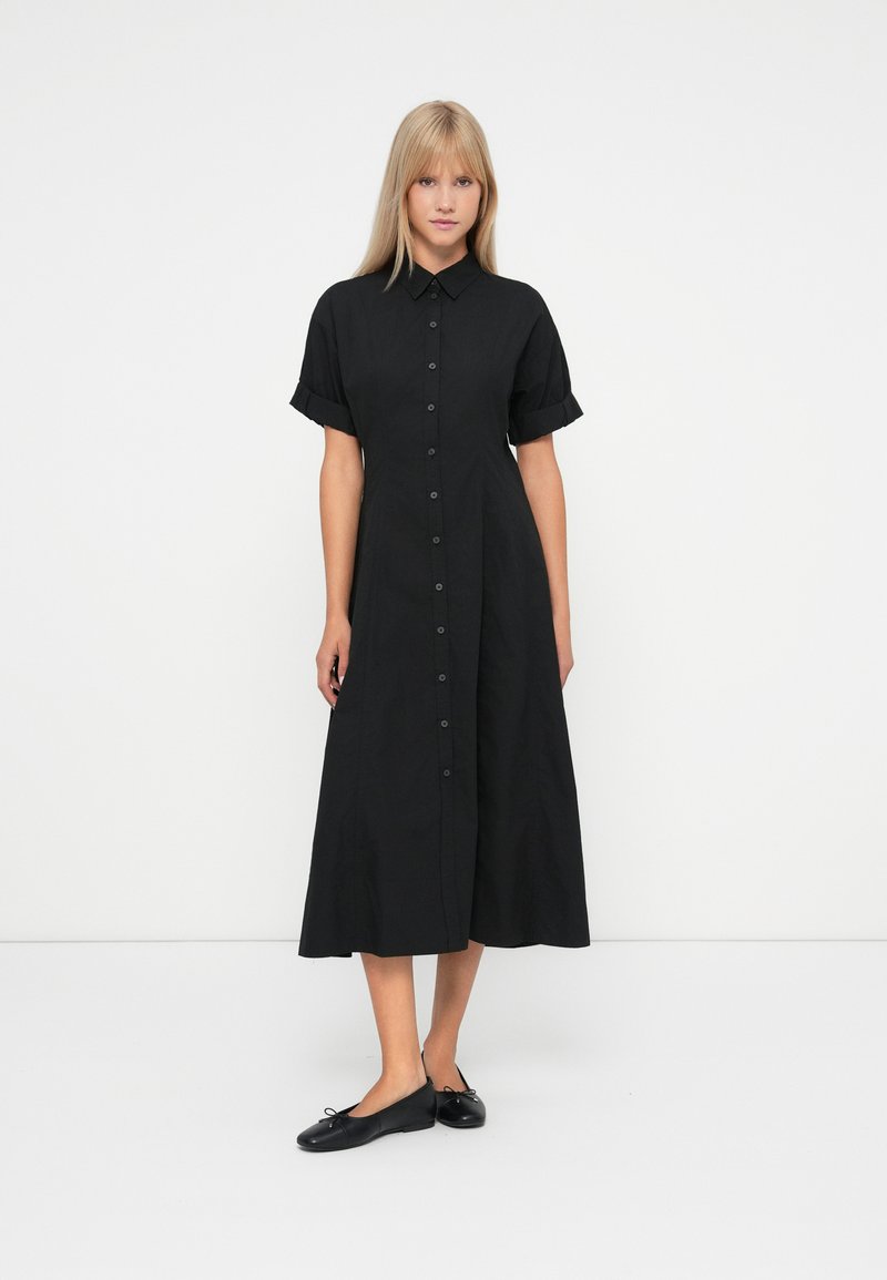 Donna in piedi con indosso un vestito midi nero a maniche corte con bottoni e colletto, abbinato a ballerine nere, contro uno sfondo bianco uniforme.