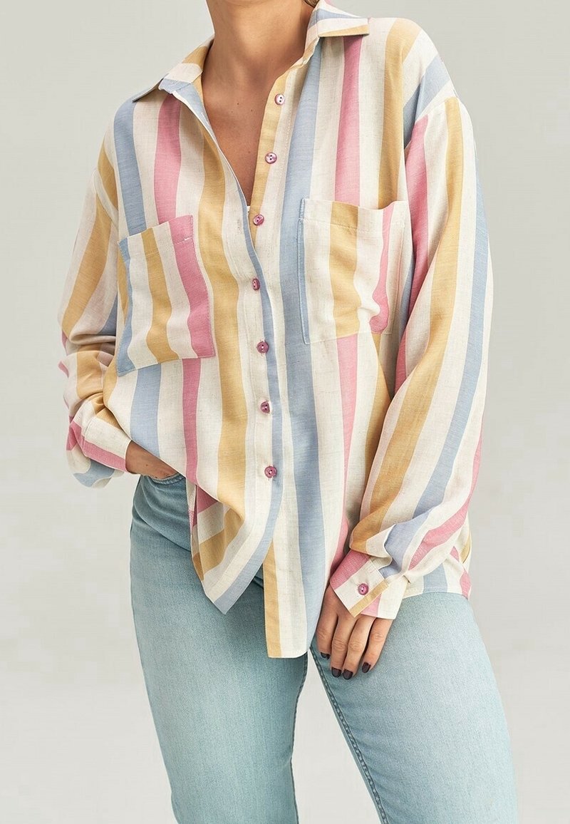 Personne portant une chemise ample boutonnée avec des rayures verticales pastel en rose, bleu, jaune et blanc, associée à un jean bleu clair.