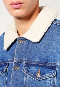 Denim takki, jossa on kerma värinen tekoshiaalinteräinen kaulus. Varustettu metallinapeilla ja rintataskuilla. Väri: keski-sininen, jossa näkyvät ompelutiedot.