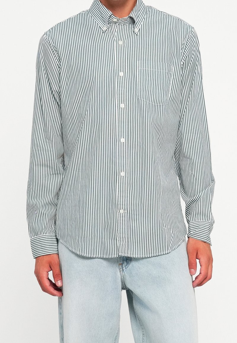 Homme portant une chemise à manches longues rayée verticalement avec une poche poitrine et un jean bleu clair.