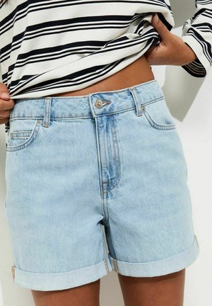 Osoba w jasnoniebieskich, podwiniętych krótkich spodenkach dżinsowych i czarno-białej, paskowanej koszuli z długim rękawem, lekko podnosząca koszulę.