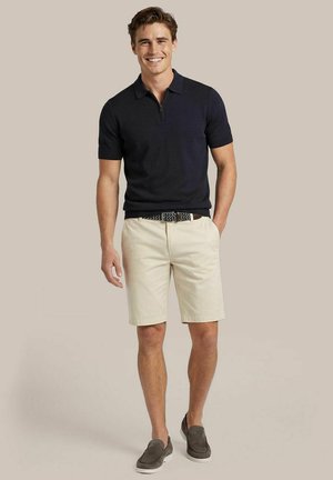 Man lacht, draagt een zwarte korte mouwen polo, beige short met een patroonriem en grijze instapschoenen, staand tegen een effen achtergrond.