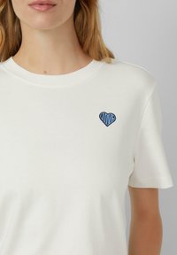 Bijela pamučna majica s kratkim rukavima, s plavim izvezenim dizajnom u obliku srca unutar kojeg se nalazi riječ "LJUBAV".