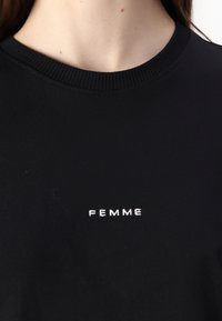 Černé bavlněné tričko s pružným výstřihem a bílým výšivkou slova "FEMME" na přední straně. Jednoduchý a moderní design.