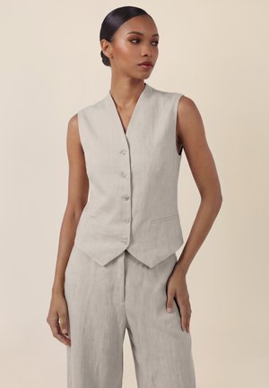 Woman wearing a sleeveless beige buttoned vest and matching wide-leg trousers, standing with one hand in pocket against a plain background.