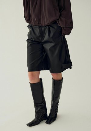 Personne portant un short en cuir noir au genou, un haut à manches longues foncé et des bottes en cuir noires montant jusqu'aux genoux, debout devant un fond uni.