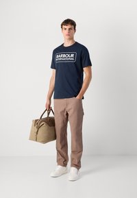 Ναυτικό βαμβακερό t-shirt με λευκό λογότυπο "BARBOUR INTERNATIONAL", συνδυασμένο με μπεζ παντελόνι και καμβαδέ σακίδιο με καφέ χερούλι. Λευκά αθλητικά παπούτσια.