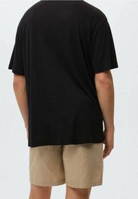 Person iført en ensfarvet sort T-shirt med korte ærmer og beige shorts, vist fra ryggen mod en ensfarvet lys baggrund.