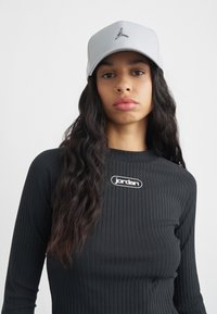 Casquette de baseball grise avec une finition texturée et un logo brodé. Chemise noire à manches longues côtelées avec un patch logo blanc sur la poitrine.