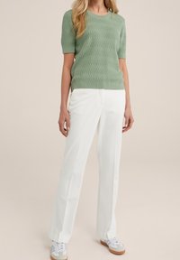 Femme portant un pull court à manches courtes vert sauge texturé, un pantalon blanc ajusté et des baskets blanches avec des semelles en caoutchouc, se tenant sur un fond neutre.