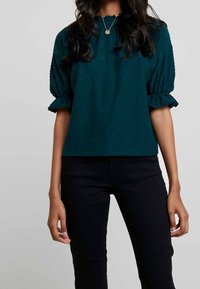 Tealfärgad blus med struktur och puffsleeves, med rund halsringning och subtila prickmönster, i kombination med svarta figurnära byxor.