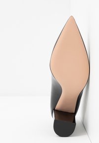 Chaussure à talon en cuir noir à bout pointu, reposant semelle vers le haut contre un mur blanc, montrant une semelle beige lisse et un talon carré.