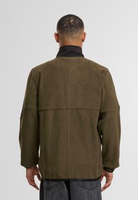 Oliivivihreä, fleece-tekstuuri takki mustalla kauluksella; sen ominaisuuksiin kuuluu pudotetut olkapäät ja suora leikkaus. Sopii arkisiin pukeutumisiin.